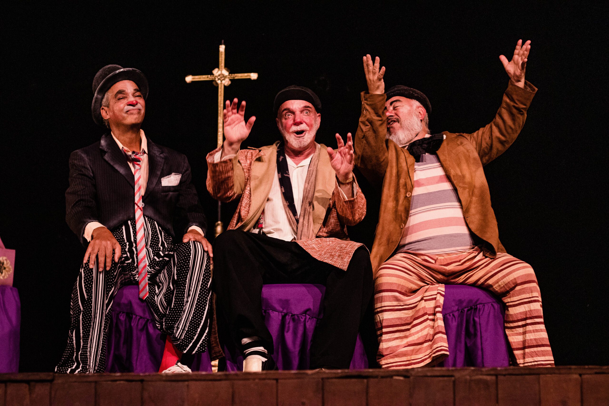 Três homens com roupas coloridas estão sentados, gesticulando e sorrindo, com uma cruz ao fundo.